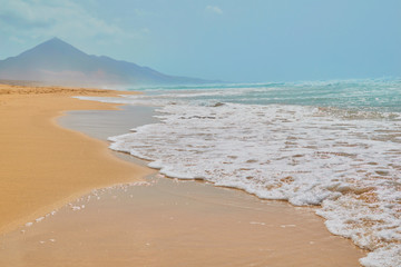 Landscape of turquoise water beach and stone mountain with golden sand and sea foam on the shore at Cofete, Fuerteventura, Canary Islands, Spain