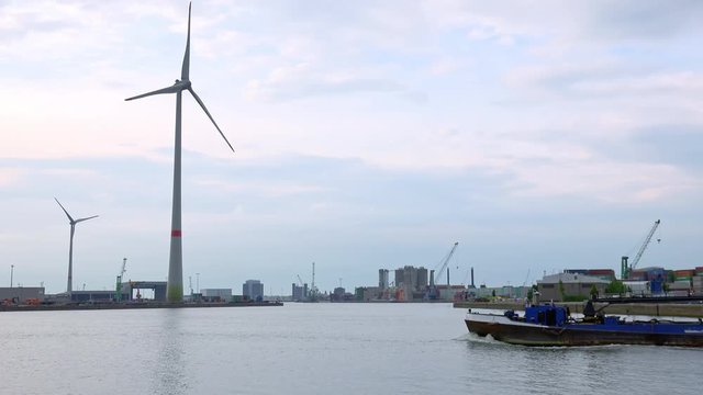 Loaded barge passing by Antwerp international seaport at  Scheldt river. Silhouettes of cranes, rotating wind turbines and stacked cargo containers near warehouse buildings on quayside. Belgium
