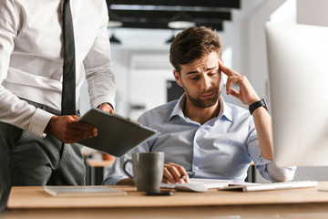 Displeased man sitting in office working with computer near his colleague who showing clipboard...