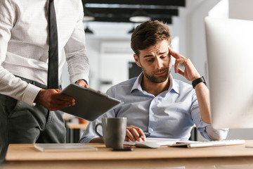 Displeased man sitting in office working with computer near his colleague who showing clipboard with documents.