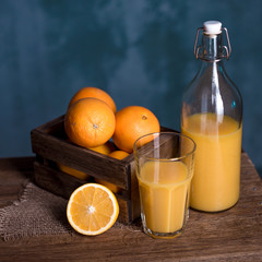 Orange juice in a transparent glass and bottle, with sliced orange, oranges in a wooden box behind, on wooden table. Square format.