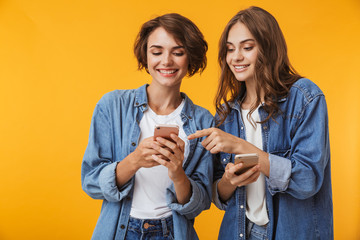 Happy emotional young women friends posing isolated over yellow background using mobile phones.