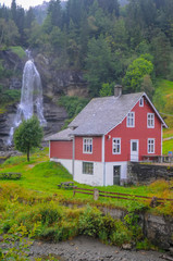 Fototapeta premium Traditional norwegian red wooden house and Steinsdalsfossen waterfall in the background, Norway