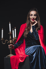 vampire in red cloak holding candelabrum and touching her fangs isolated on black