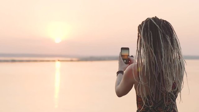 attractive hippie woman with dreadlocks making photo on smart phone at sunset near sea