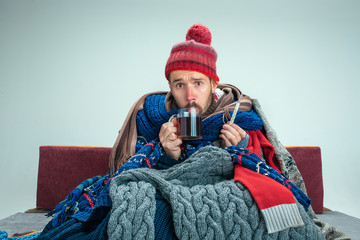 Bearded sick man with flue sitting on sofa at home or studio with cup of tea covered with knitted...