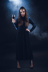 grim vampire holding wineglass with blood on dark background with smoke