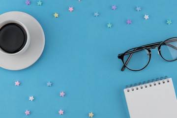 A cup of coffee, blank notebook and eyeglasses decorate with pastel star pattern on blue background with copy space