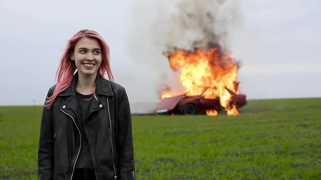 Young beautiful girl with pink hair and in a leather jacket stands on the background of an exploding car, a hipster and a burning car, car explosion