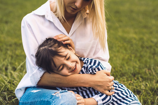 Image of happy cute little girl embrace and lying on her mother legs outdoor. Loving mother and daughter sitting on green grass in the park. Mom and kid cuddling, have fun outside. Good relationship