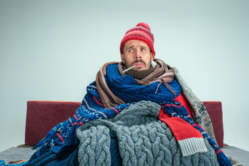 Bearded sick man with flue sitting on sofa at home or studio with thermometer covered with knitted...
