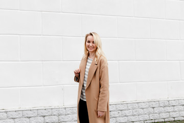 Beautiful young stylish blonde woman wearing long beige coat posing near white street wall. Trendy casual outfit. Street fashion.