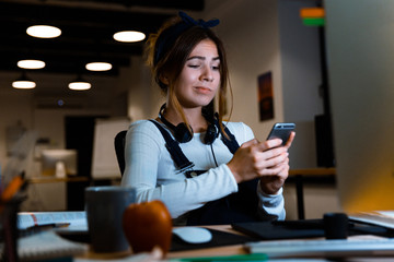 Confused young woman designer sitting in office working using mobile phone at night.