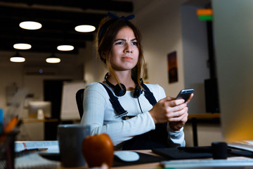 Confused young woman designer sitting in office working using mobile phone at night.