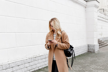 Beautiful young stylish blonde woman wearing long beige coat and black backpack walking through the city streets with smartphone. Trendy casual outfit. Street fashion. Technology, communication.