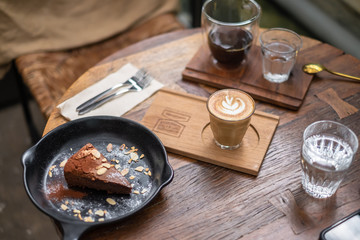 Coffee latte art in glass cup with a piece of chocolate cake and hot drip coffee , forks and wooden heart on wooden table background. Coffee lover concept.