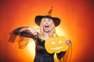 Beautiful young surprised woman in witches hat and costume pointing hand - showing products....