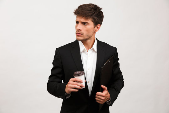 Serious businessman standing isolated holding clipboard looking aside drinking coffee.