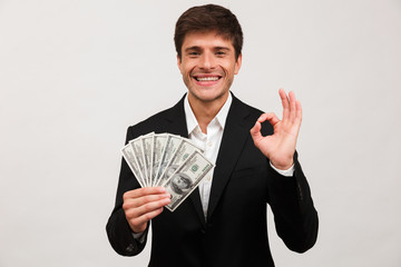 Happy businessman standing isolated holding money make okay gesture.