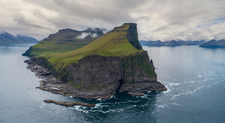 AERIAL: Tiny white lighthouse close to black cliff looks over the tranquil sea.