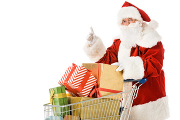 santa claus with shopping cart full of gift boxes pointing up isolated on white