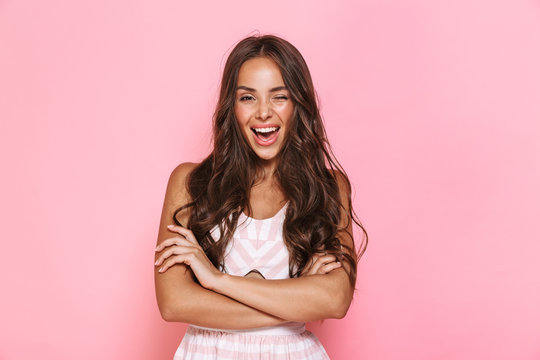 Image of cute woman 20s with long hair wearing dress smiling at camera with arms crossed, isolated over pink background