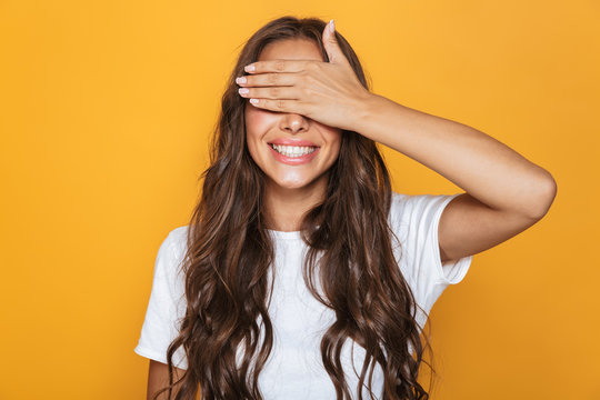 Image of excited woman 20s with long hair smiling and covering eyes with hands, isolated over yellow background