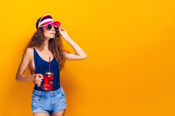 Sensual young woman in summer outfit, cap, sunglasses holding jar with fresh beverage and while standing on bright yellow background