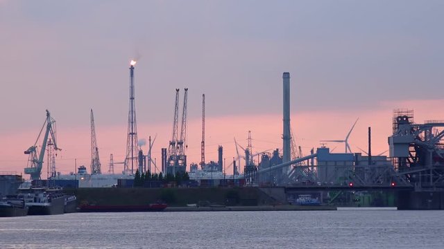Futuristic scenery with silhouettes of cranes and rotating wind turbines at marine cargo terminal. Gas flare against beautiful sunset sky on background. Seaport area at evening. Antwerp, Belgium.