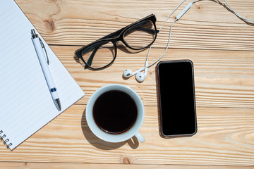 Cup of black coffee and smart phone with office supplies; pen, notebook and eyes glasses on blue wooden table background. Top view with copy space