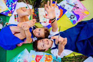 Authentic artist children in blue aprons girl and boy paints tickle each other by paint brushes, lying in creative chaos palette watercolor paints, brush, drawings.Painting outdoor. Top view © Iryna