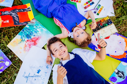 Authentic artist children in blue aprons girl and boy paints tickle each other by paint brushes, lying in creative chaos palette watercolor paints, brush, drawings.Painting outdoor. Top view