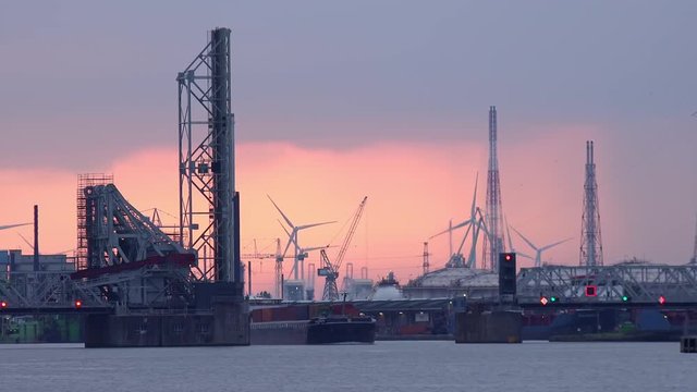 Loaded barge passing by raised drawbridge over Scheldt river. Silhouettes of cranes and rotating wind turbines view against sunset sky. Antwerp international seaport, Belgium. Industrial scene