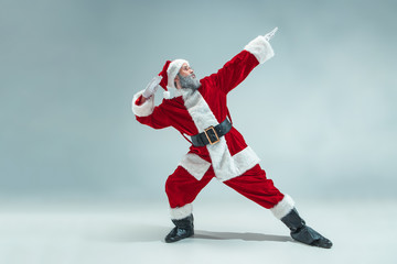 Fototapeta na wymiar Funny serious guy with christmas hat dancing at studio. New Year Holiday. Christmas, x-mas, winter, gifts concept. Man wearing Santa Claus costume on gray. Copy space. Winter sales.