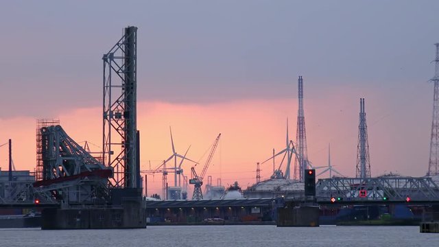 Beautiful evening view of raised truss bridge over Scheldt river, cranes, buildings of Antwerp seaport and rotating wind turbines against amazing sunset sky. Belgium. Abstract industrial scenery