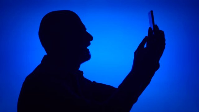 Silhouette of senior man using mobile phone on blue background. Male's face in profile doing selfie on cellphone. Black contur shadow of grandfather's half-face making photos on smart phone