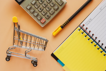 Mini shopping cart or trolley with opened spiral notebook, pen and calculator on beige color background for spending plan concept