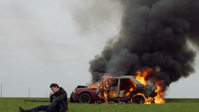 A guy lights a cigarette against the backdrop of his burning car, the harm of smoking, a man smokes against the background of a fire car, slow-mo