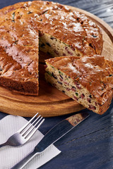 Appetizing unsweetened pie with a salty filling of olives, sausages, meat, vegetables, peas. Big pie lies on a beautiful wooden round board. Cutlery lies on a white paper napkin.