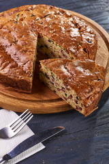 Appetizing unsweetened pie with a salty filling of olives, sausages, meat, vegetables, peas. Big pie lies on a beautiful wooden round board. Cutlery lies on a white paper napkin.