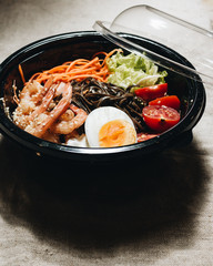 Delicious meal of different ingredients, egg, shrimps, tomatoes, carrot, lettuce, sesame seeds and seaweed in black plate, Asian cuisine