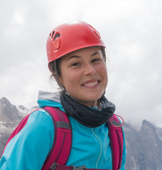 portrait of a young and beautiful female mountain climber in a blue jacket and red helmet smiling with a mountain landscape background