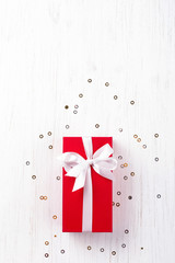Fototapeta na wymiar Christmas gift wrapped in red paper tied with white ribbon, decorative paper roll and candy cane lollipop on white wooden background with sparkles. Flat lay, copy space.