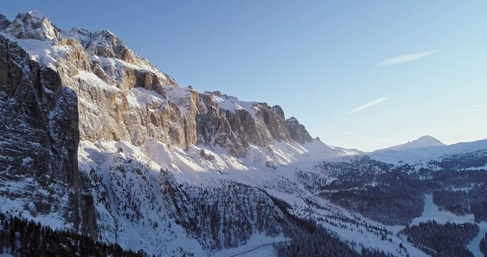 Going down aerial along snowy alpine steep rocky cliff valley with woods forest.Sunset or sunrise, clear sky.Winter Dolomites Italian Alps mountains outdoor nature establisher.4k drone flight