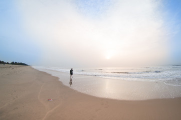Man standing watching the sunrise on a beach in pondicherry chennai, with the waves crashing in and the clouds glowing pink with the light. A perfect vacation shot for an indian vacation