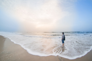 Fototapeta na wymiar Slow shutter shot of man standing on beach in chennai pondicherry watching sunrise on a cloudy day with the waves crashing around