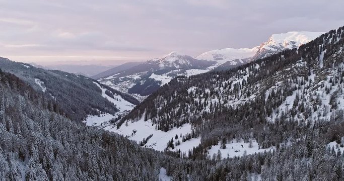 Backward aerial to snowy valley with woods forest at Sella pass.Sunset or sunrise,cloudy sky.Winter Dolomites Italian Alps mountains outdoor nature establisher.4k drone flight