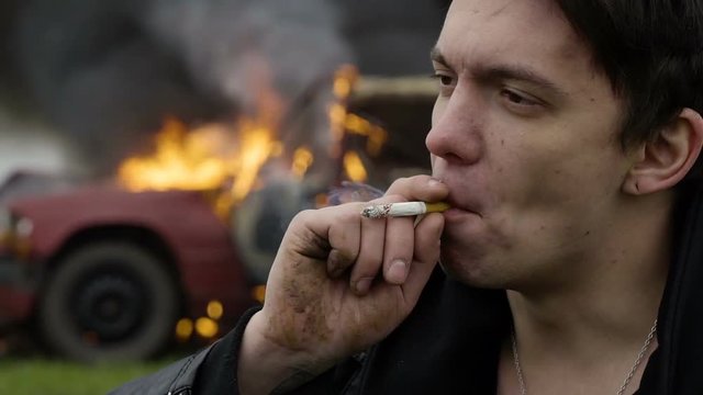 The thoughtful guy smokes against the background of his burning car, melancholy, an explosion of a car