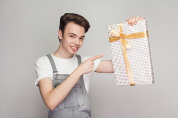 Fototapeta na wymiar Cheerful modern teenager in demin overalls and white t-shirt standing and pointing finger to present with yellow bow and toothy smile, looking at camera. Indoor, isolated, studio shot, grey background