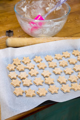 Ready to bake biscuits: cookie dough in star shapes on baking sheet, with pink spatula, mixing bowl, cutter and rolling pin on messy kitchen bench